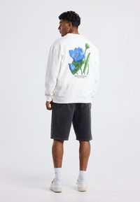 Valkoinen huppari, jossa on suuri värikäs kukkakuva selkäpuolella, yhdistetty mustiin shortsien ja valkoisten tennareiden kanssa. Vähäistä tekstuuria, rento malli.