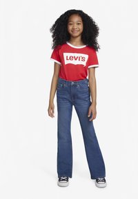 Rotes T-Shirt mit weißen Abschlüssen und weißem Levi's-Logo, kombiniert mit hoch taillierten blauen Schlagjeans und schwarzen Turnschuhen.
