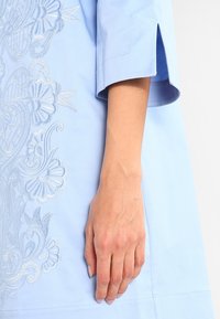 Gros plan d’une main reposant à côté d’une robe bleu clair avec une broderie florale complexe et une manche courte fendue.