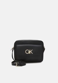 Borsa a tracolla in pelle nera con tasca anteriore a patta e logo CGK oro, tracolla regolabile e chiusura con zip su sfondo bianco.