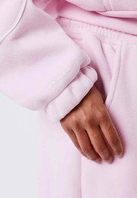 Sweat-shirt rose clair avec poignets élastiques, présentant une texture douce et un logo "PXP" blanc sur la manche, associé à un jogging assorti.