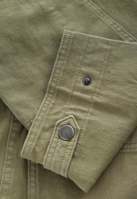 Gros plan sur le poignet en tissu kaki d'une chemise avec deux boutons-pression et des coutures visibles.