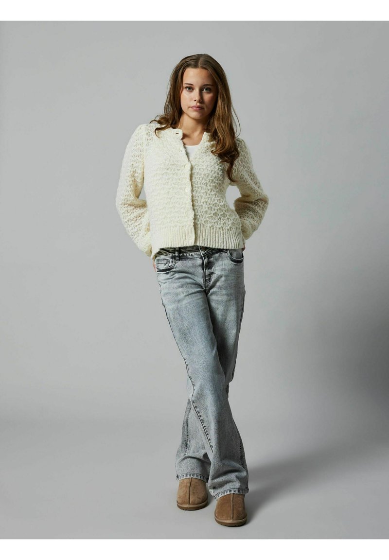 Cardigan in maglia di crema con texture a trafori, vita aderente e chiusura con bottoni, abbinato a jeans denim chiari e stivali color nocciola. Sfondo grigio.
