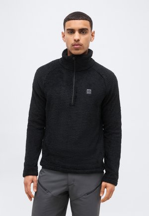 Jeune homme portant une polaire noire texturée à demi-fermeture éclair avec un logo 66 North, associée à un pantalon gris, debout devant un fond uni.