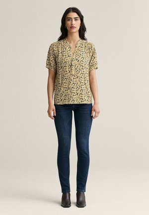 Nainen, jolla on päällään keltainen kukallinen lyhythihainen paita, tummansiniset kapeat farkut ja tummanruskeat nilkkurit, seisoo yksinkertaisen taustan edessä.