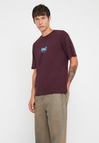 Burgundy cotton t-shirt with a blue logo print, relaxed fit, and short sleeves, paired with light beige denim pants. Simple design, no patterns.