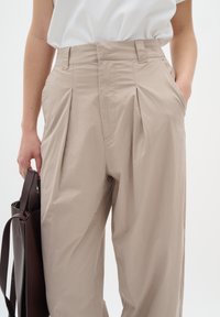 Světle béžové plisované kalhoty vyrobené z lehkého materiálu, s vysokým pasem a bočními kapsami, doplněné hnědou kabelkou.