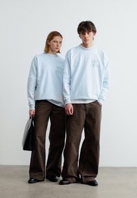 Δύο άτομα φορούν ανοιχτό μπλε φούτερ με την επιγραφή "EST 1978", καφέ παντελόνια φαρδιάς γραμμής και μαύρα παπούτσια, στέκονται μπροστά σε ένα απλό λευκό φόντο.