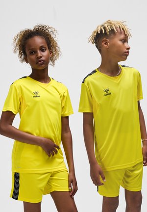 To børn iført gule hummel sportstrøjer og matchende shorts, står mod en ensfarvet lys baggrund.