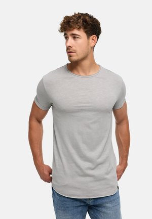 Młody mężczyzna z kręconymi włosami, ubrany w dopasowany jasnoszary t-shirt i niebieskie jeansy, patrzy lekko w lewo, trzymając ręce przy bokach.