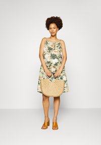 Vestido de verão em tecido suave com um padrão floral verde, apresentando um laço na frente. Segura uma bolsa redonda de palha trançada e usa sandálias de salto castanhas.