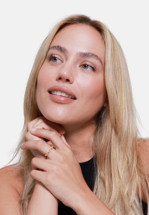 Goldring mit Kettenglieddesign, der an den Fingern getragen wird, mit einer glatten Oberfläche. Mittellange blonde Haare, natürliches Makeup und schwarzes Oberteil sichtbar.