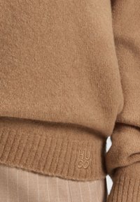 Brauner Strickpullover mit gerippten Bündchen und Saum, auf dem ein reliefartiges "HUGO"-Logo prangt. Weiche Textur, schlichtes Design und lockere Passform.