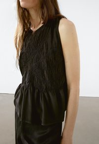 Czarny top bez rękawów z teksturowanymi przeszyciami na klatce piersiowej oraz zwiewnym dołem w stylu peplum, wykonany z lekkiego materiału.