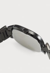 Orologio da polso in metallo nero con cassa liscia e arrotondata, quadrante circolare piatto e corona laterale. Caratterizzato da un cinturino elegante con accenti lucidi.