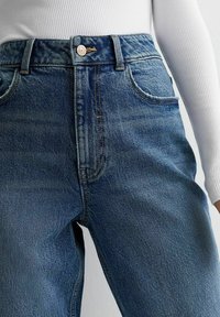 Jeans in denim blu con vita alta, design a cinque tasche e bottoni in ottone. Il tessuto ha una texture liscia con una leggera elasticità.