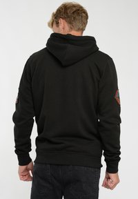 Zwarte hooded sweatshirt met een gestructureerde afwerking, voorzien van een patch op de linkermouw en een relaxte pasvorm, weergegeven vanuit de achterkant.