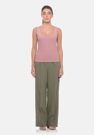 Donna in piedi di fronte, con un top rosa senza maniche, pantaloni larghi verde oliva e sandali bianchi su uno sfondo bianco semplice.