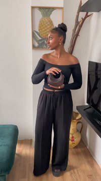 Top nero con spalle scoperte e pantaloni larghi neri; tiene in mano una piccola borsa curvata marrone. Lo sfondo presenta un'illustrazione di un ananas dorato.