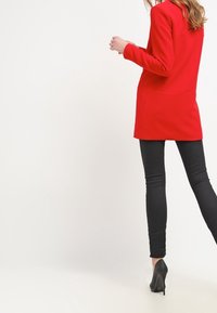 Roter Blazer mit taillierter Passform, langen Ärmeln und einem Ein-Knopf-Verschluss, kombiniert mit schwarzen Skinny-Jeans und spitzen schwarzen High Heels.