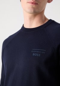 Temno modra pletena majica s krojem za vrat. Ima diskreten logotip Porsche in svetlejše modro besedilo "BOSS" na prsih.
