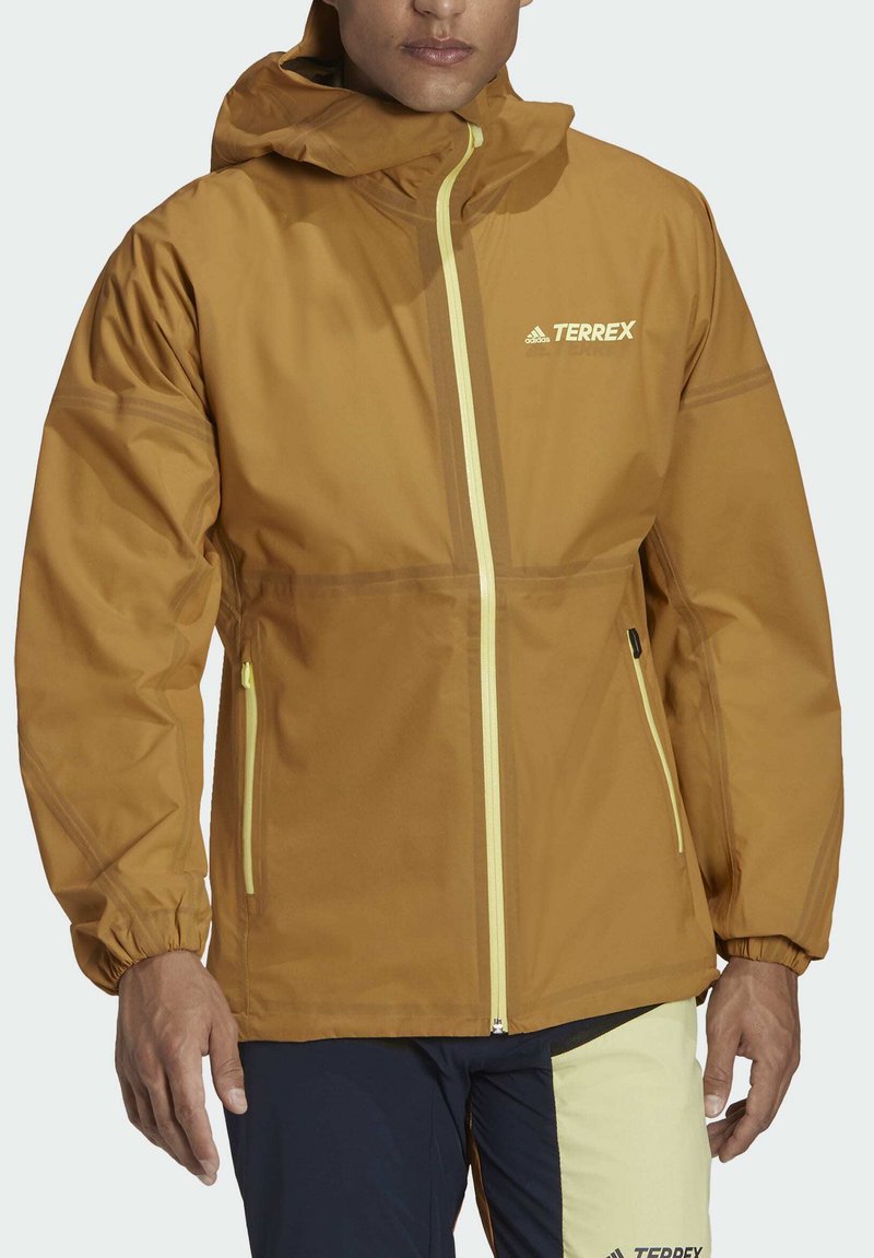 Chaqueta impermeable marrón con capucha, cremalleras amarillas, puños elásticos y logotipo. El material parece suave con un acabado mate.