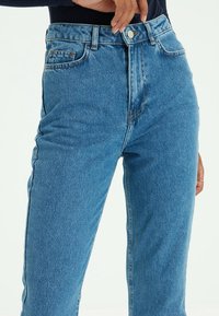 Jean en denim bleu taille haute avec une coupe classique, doté d'un design à cinq poches et d'un léger effet délavé. Fermeture à boutons à la taille.