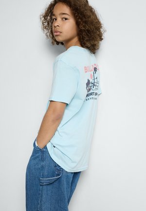 T-shirt en coton bleu clair avec un design graphique dans le dos. Porté avec un jean en denim bleu, offrant une coupe décontractée et des poches.