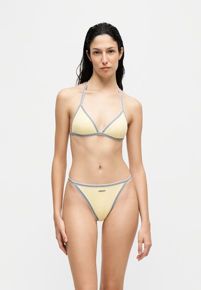 Femme aux longs cheveux foncés portant un bikini Versace jaune pâle avec des bordures noires, debout devant un fond blanc uni.