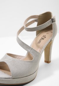 Sandale à talons hauts gris clair en daim souple avec des brides croisées à la cheville, bout ouvert et talon épais. Finition intérieure lisse.