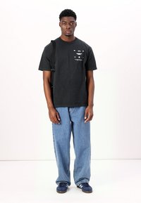 Black short-sleeve T-shirt with graphic design on the chest, paired with light blue loose-fitting jeans and navy sneakers. Simple strap across shoulder.