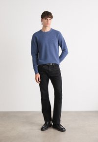 Blå genser med rund hals, ribbestrikket kant, brukt over svarte straight-leg jeans og svarte sko, satt mot en minimalistisk bakgrunn.