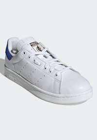 Bílé kožené tenisky s plochou podrážkou, modrým akcentem na patě a perforovaným detailem na boční straně. Obsahují kulaté tkaničky a štítek na jazyku.