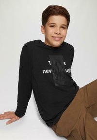 Zwarte lange mouwen shirt met witte tekst "Er is nooit genoeg tijd" op de voorkant, gecombineerd met een bruine broek. Gladde textuur, casual ontwerp.