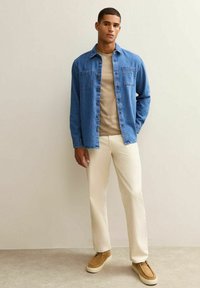Lichtblauwe denimshirt met twee borstzakken, beige t-shirt, crèmekleurige broek en lichtbruine instap schoenen. Model staat tegen een neutrale muur.
