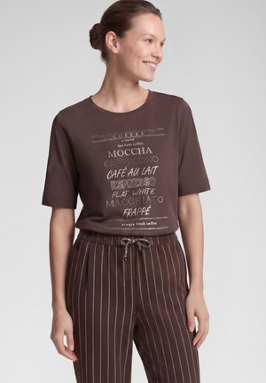 Woman wearing a brown t-shirt with coffee drink names and brown pinstriped pants, smiling and looking to the side.