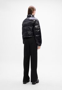 Giacca a vento cropped nera con finitura lucida, caratterizzata da sezioni trapuntate orizzontali, colletto alto e polsini elasticizzati, indossata con pantaloni neri.