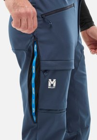 Pantalons softshell bleu marine avec une fermeture éclair sur le côté. Comprend une doublure en mesh, des poches zippées et un logo Millet blanc bien visible sur la cuisse.