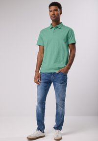 Camisa polo de algodón verde con cuello, combinada con jeans azules desgastados y zapatillas blancas. El modelo está posando, con las manos en los bolsillos.