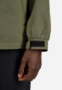 Olive roheline jakk, millel on lukuga esiosa ja elastsed mansetid. Velcro aktsendid mansetil. Sile, tekstureeritud kangas. Suurendus pildil, kus käsi hoiab jakki.