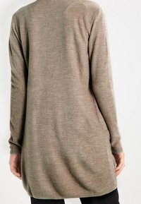 Długi, dziergany kardigan w jasnobrązowym odcieniu. Posiada prążkowaną teksturę i zaokrąglony dół, długie rękawy oraz luźny krój.