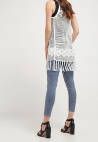 Weißes Netz-Top mit Häkel-Details und Fransen am Saum, kombiniert mit blauen Skinny-Jeans und schwarzen Absatzsandalen.