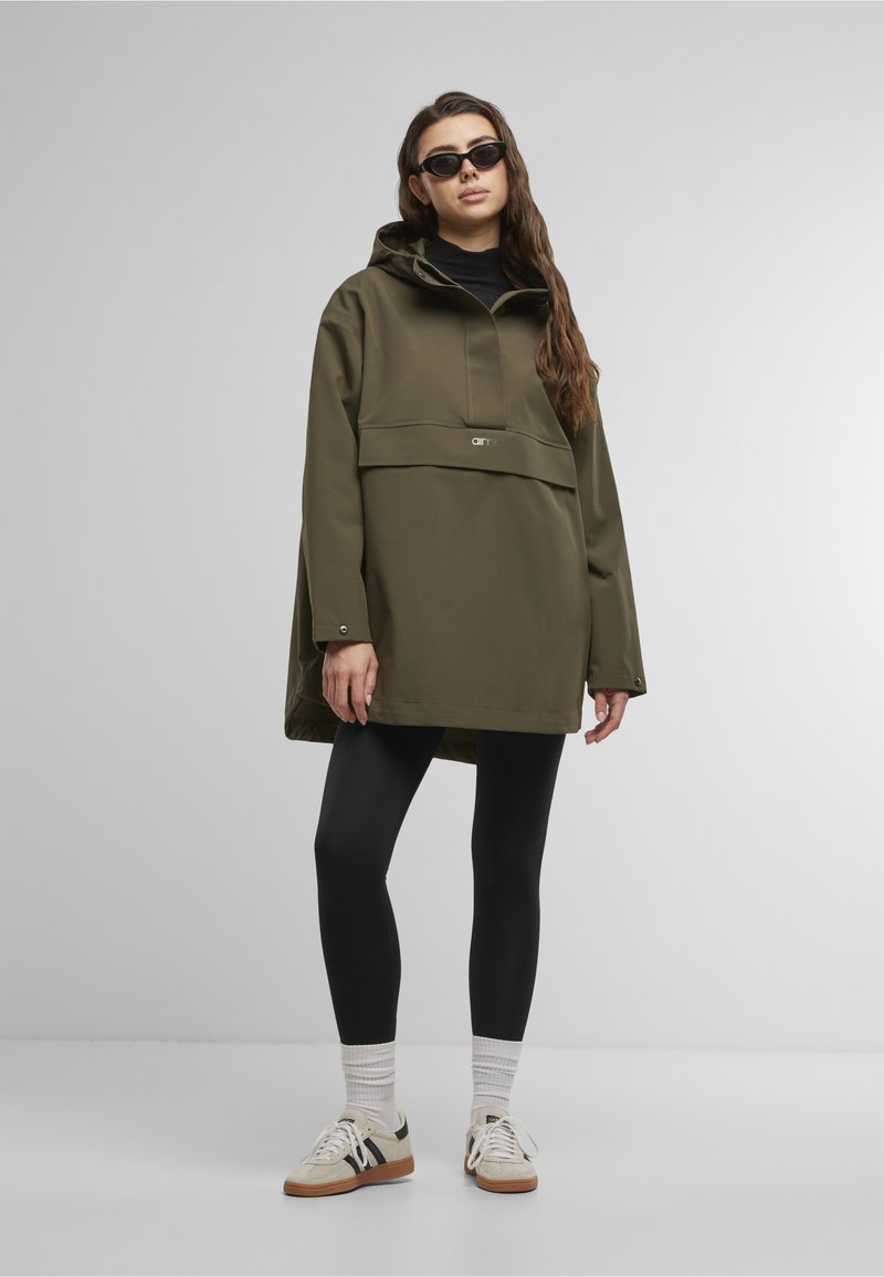 Donna che indossa una giacca con cappuccio oversize verde oliva, leggings neri, calzini bianchi, sneakers e occhiali da sole neri, in piedi su uno sfondo semplice.