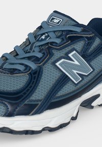 Μπλε και ναυτικό αθλητικό παπούτσι New Balance με δίχτυ, παχύ λευκό πέλμα και ανοιχτογάλα κορδόνια δεμένα με φιόγκο.