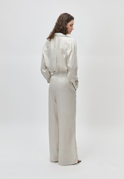 Γυναίκα με σγουρά μαλλιά που φοράει ανοιχτόχρωμο πουκάμισο με λεπτές ρίγες και παντελόνι με φαρδιά πόδια που ταιριάζουν, στέκεται πλαγίως.