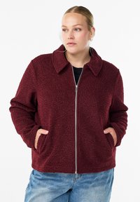 Veste en polaire bordeaux avec un col, fermeture éclair à l'avant et deux poches latérales. Texture douce et coupe décontractée. Portée par-dessus un haut noir.
