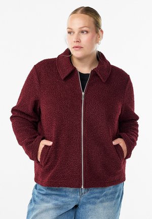 Bordeaux fleece jack met een kraag, voorsluiting met rits en twee zijzakken. Zachte textuur en een ontspannen pasvorm. Draagt over een zwart top.