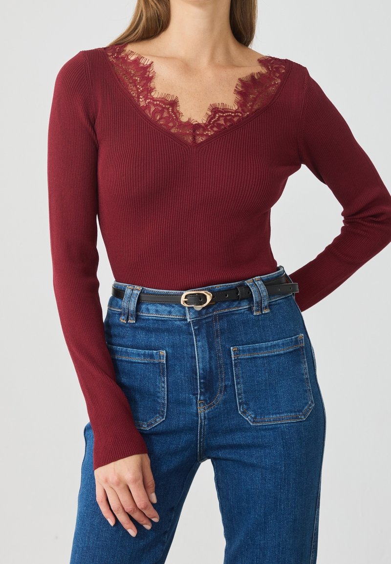 Femme portant un haut côtelé bordeaux à manches longues avec un détail en dentelle au niveau du col et un jean bleu taille haute avec des poches avant et une ceinture noire.