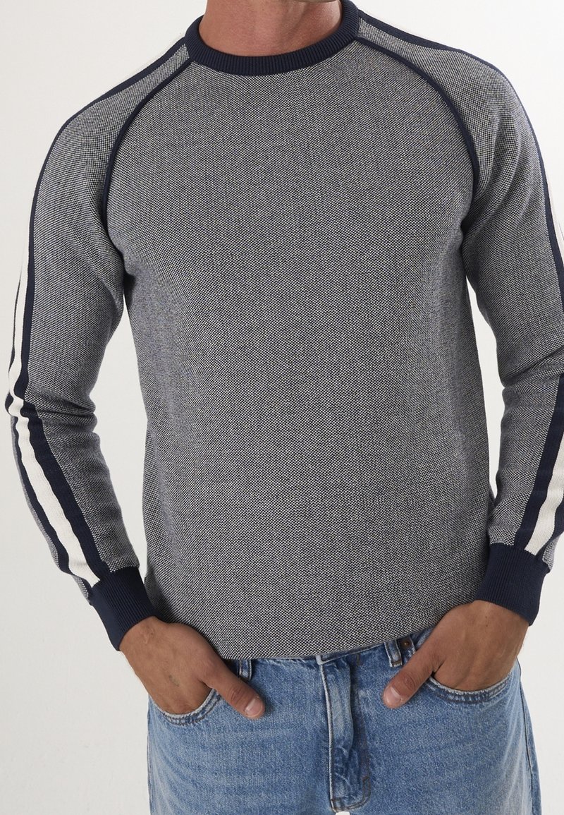 Homme portant un pull gris texturé avec des rayures bleu marine et blanches sur les manches, associé à un jean bleu, mains dans les poches.
