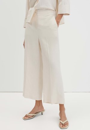 Donna che indossa pantaloni corti larghi color crema e sandali abbinati con tacco basso e punta aperta, in piedi davanti a uno sfondo bianco semplice.